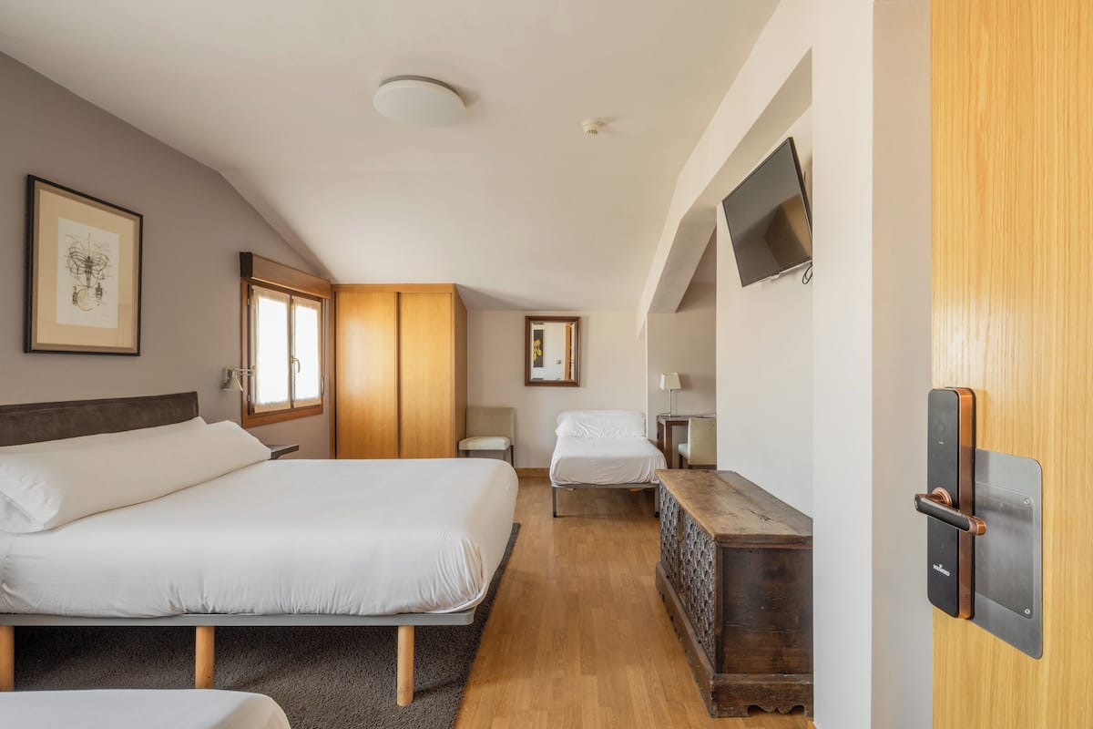 Een slaapkamer met een groot bed, een kleiner bed, een houten dressoir, een aan de muur bevestigde televisie en een inlijstbaar kunstwerk.