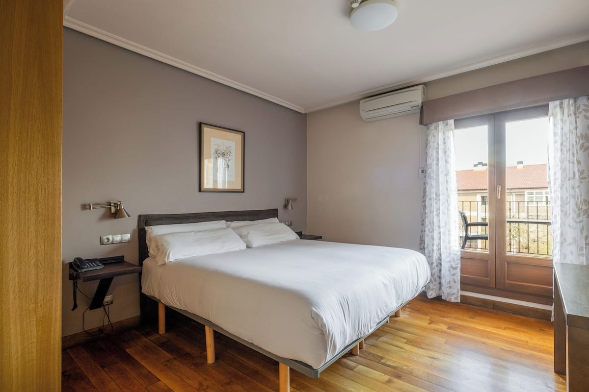 Een slaapkamer met een bed, nachtkastje, lamp, airconditioning en een raam met gordijnen.