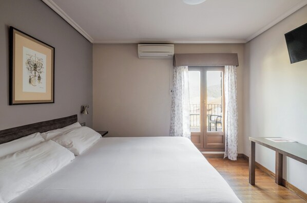 A hotel room with a large bed, a desk, a framed picture on the wall, an air conditioning unit, and a view of the outdoors through a window. - Hostal Izar-Ondo (Arbizu)