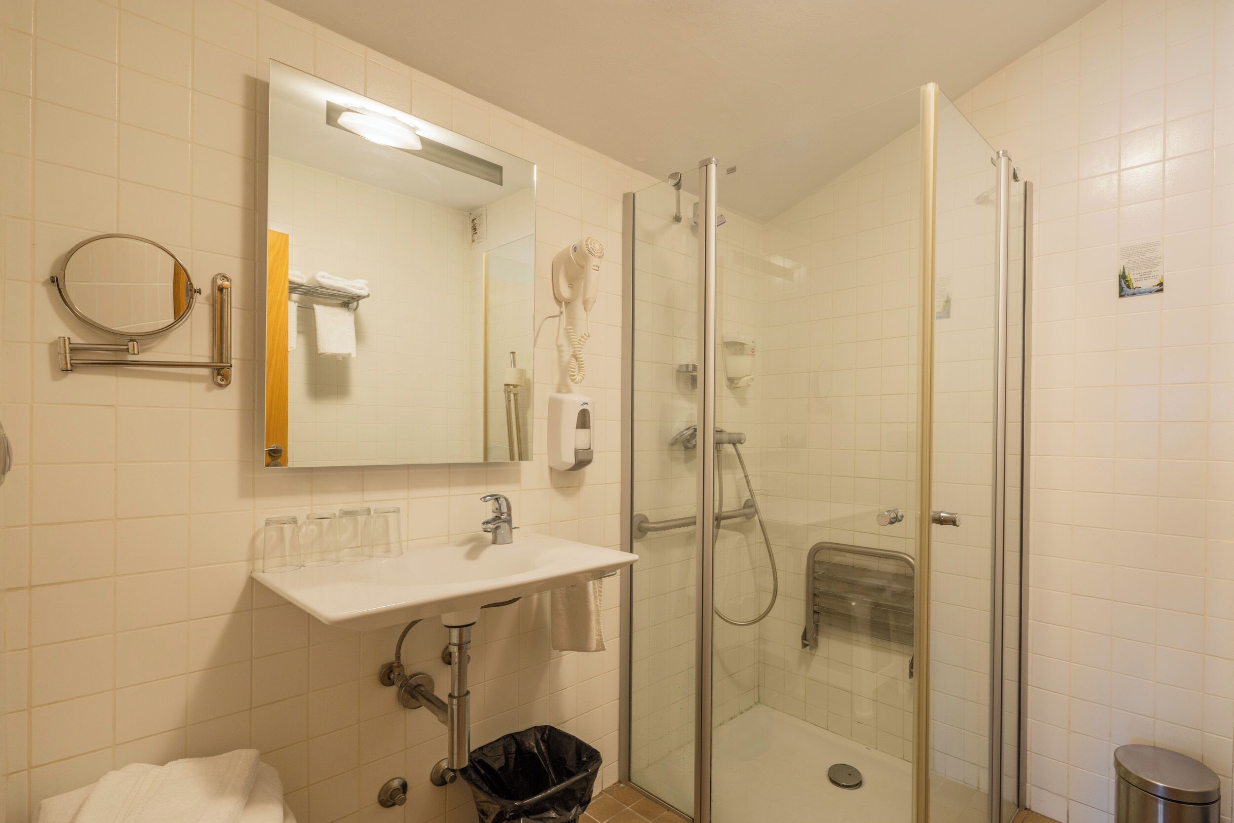 Un baño moderno con ducha envuelta en vidrio, un lavabo blanco con espejo arriba y un bote de basura negro.