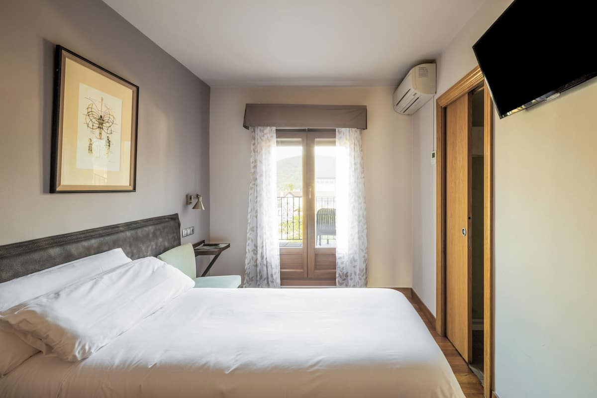 Een hotelkamer met een bed, een ingelijste foto aan de muur, een raam met gordijnen, een airconditioning en een televisie die aan de muur hangt.