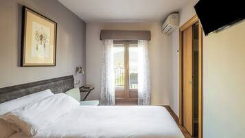 A hotel room with a bed, a framed picture on the wall, a window with curtains, an air conditioning unit, and a television mounted on the wall.