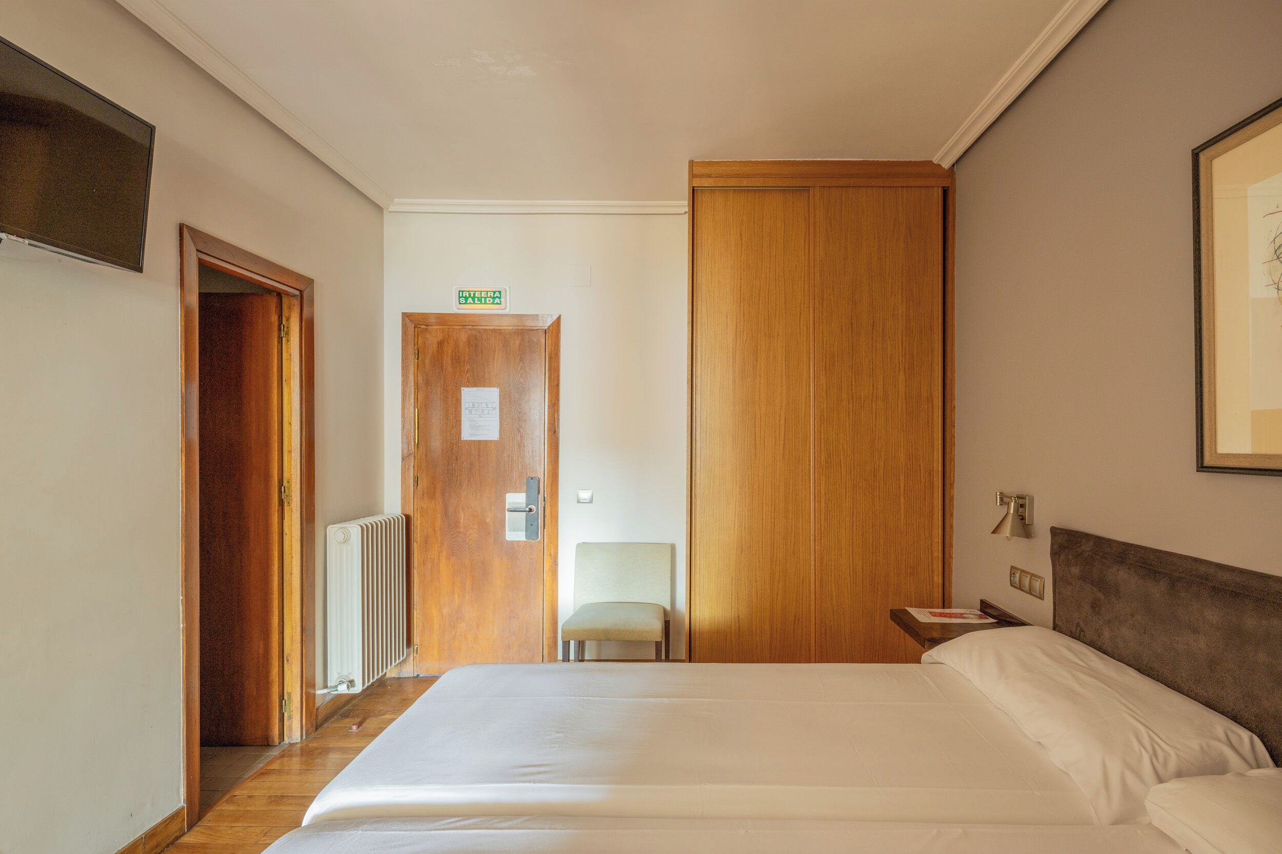Een hotelkamer met een groot bed, een houten kledingkast, een stoel en een tafeltje met een lamp.