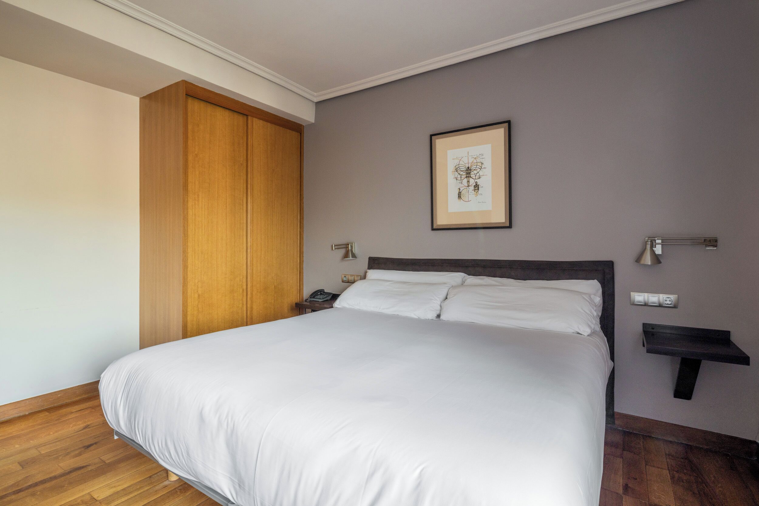 Una cama bien tendida con sábanas blancas, un armario de madera, una obra de arte enmarcada en la pared y una mesita de noche con una lámpara.