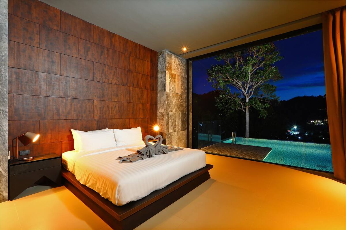A modern bedroom with a large bed, a nightstand, and a view of a pool at night.