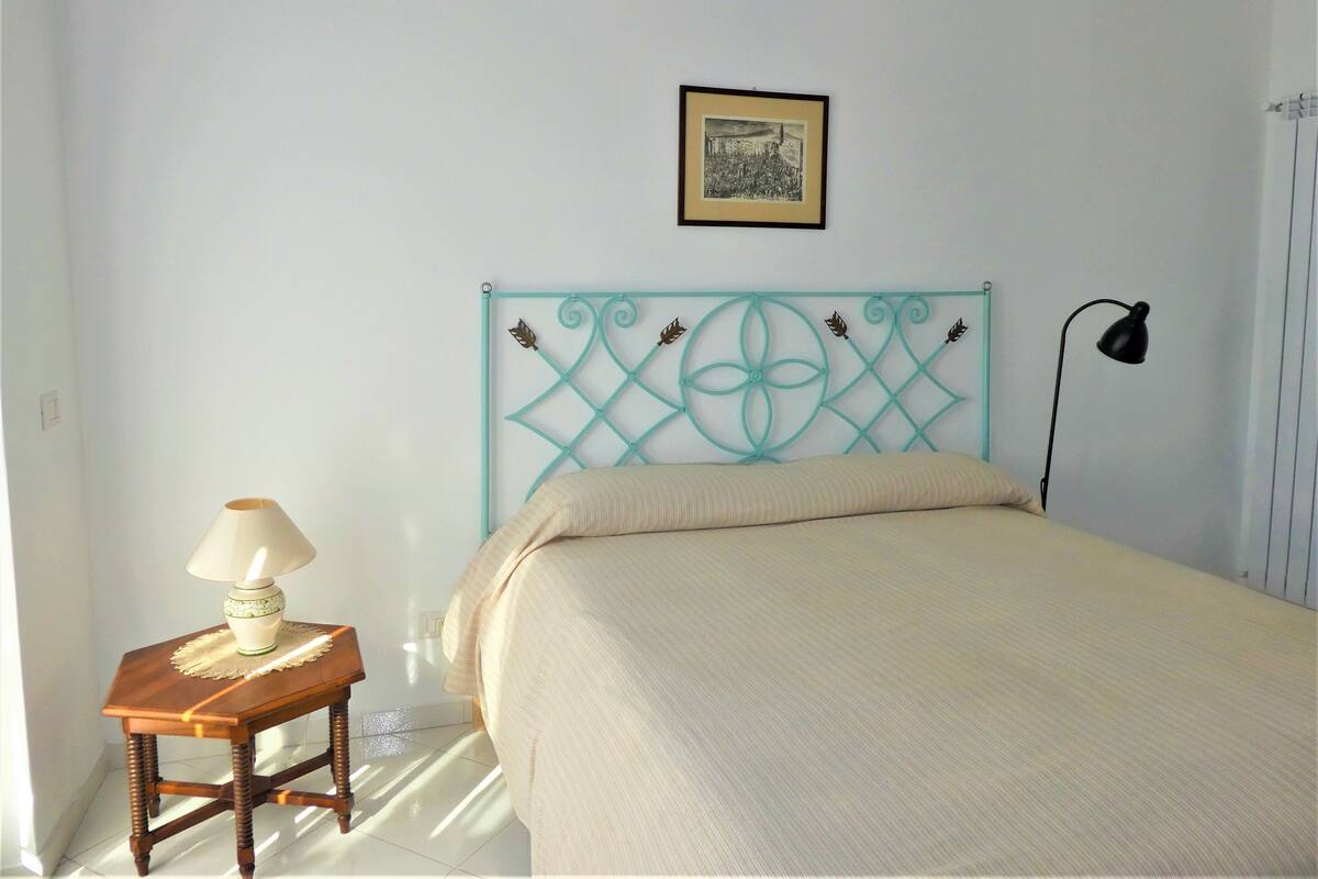 A bedroom with a bed, a nightstand with a lamp, a framed picture on the wall, and a window with blinds.