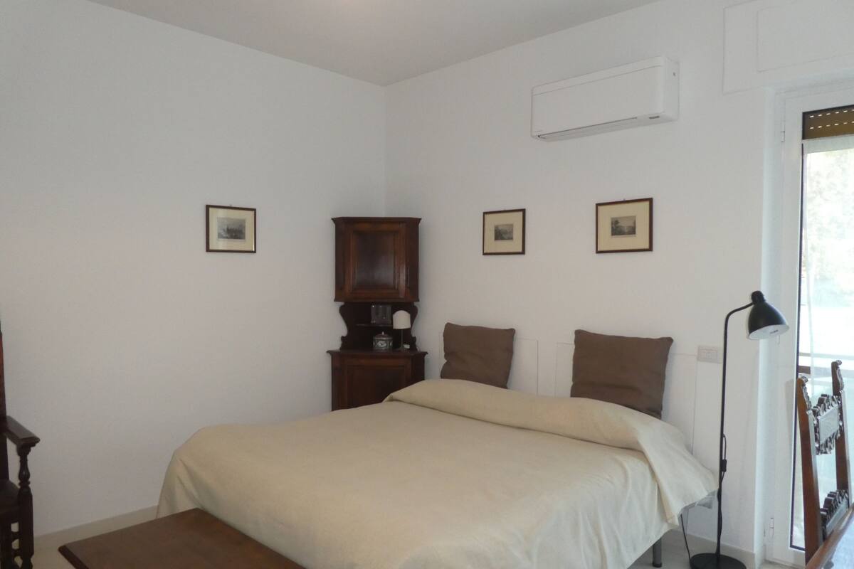 A bedroom with a bed, a wooden nightstand, a desk, and a lamp. There are three framed pictures on the wall.
