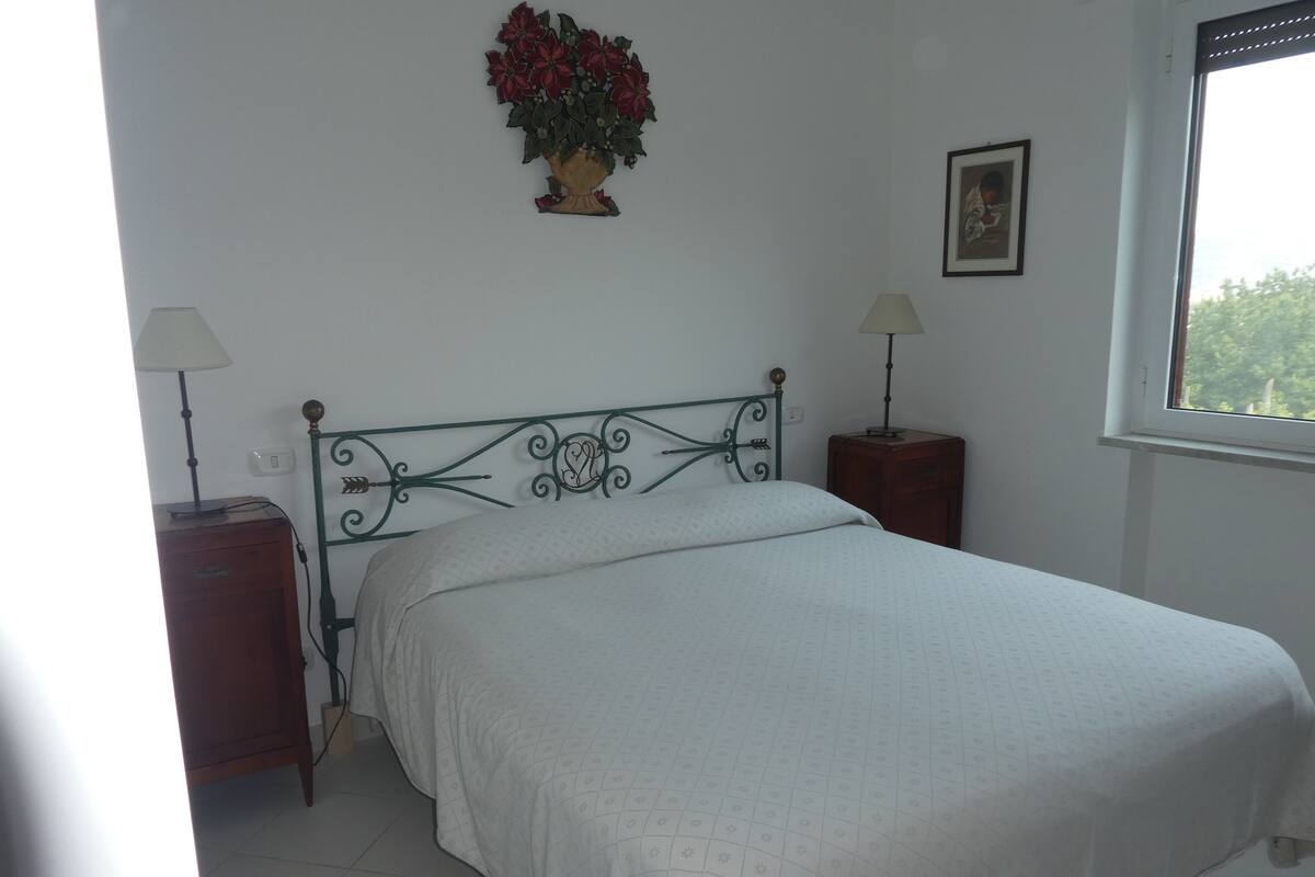 A bedroom with a bed, two bedside tables, a nightstand, a wall-mounted lamp, a framed picture, and a window with a view.