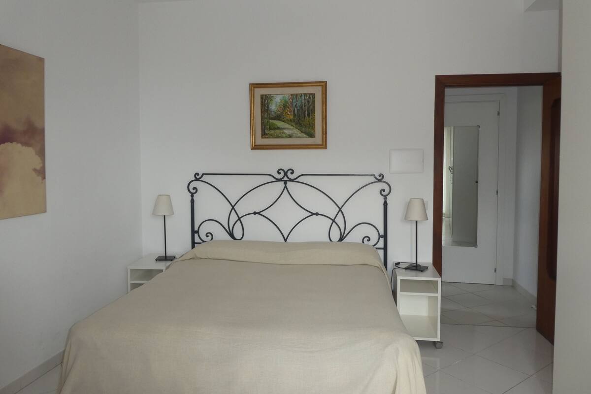 A bedroom with a bed, two bedside tables, a nightstand, a painting on the wall, and a door leading to another room.