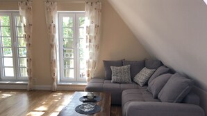 Un salon moderne avec un canapé gris, une table basse en bois et une vue sur la verdure grâce à de grandes fenêtres ornées de rideaux à motifs.