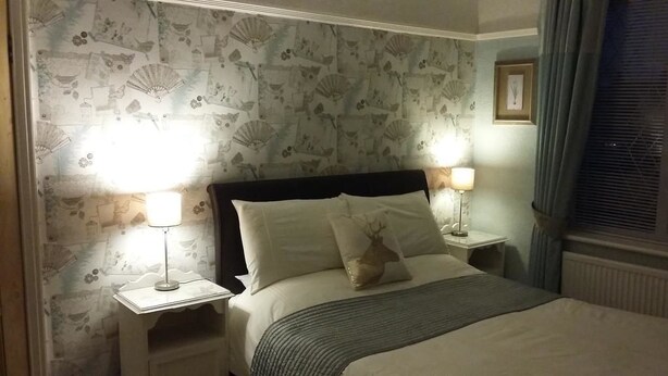 A neatly made bed with white linens and two decorative pillows, flanked by bedside tables with lamps. The room features a patterned wallpaper and a framed picture on the wall. - EsJays at Knowles House (Lytham St. Anne's)