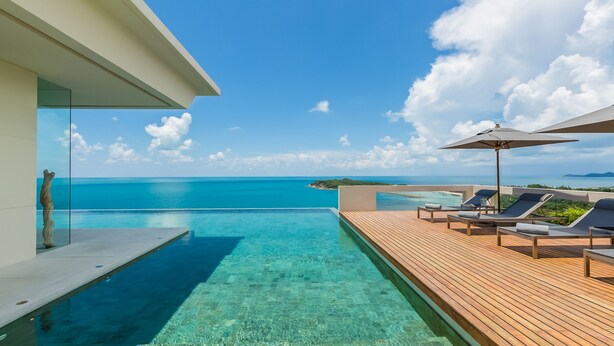Un ciel d’un bleu éclatant, une terrasse en bois avec des chaises longues et des parasols, et une piscine donnant sur l’océan.