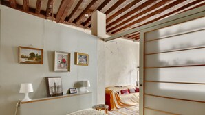 Camera da letto con un letto, lampade da comodino, un tavolino e una sedia. Alle pareti ci sono quadri incorniciati e una finestra con persiane.