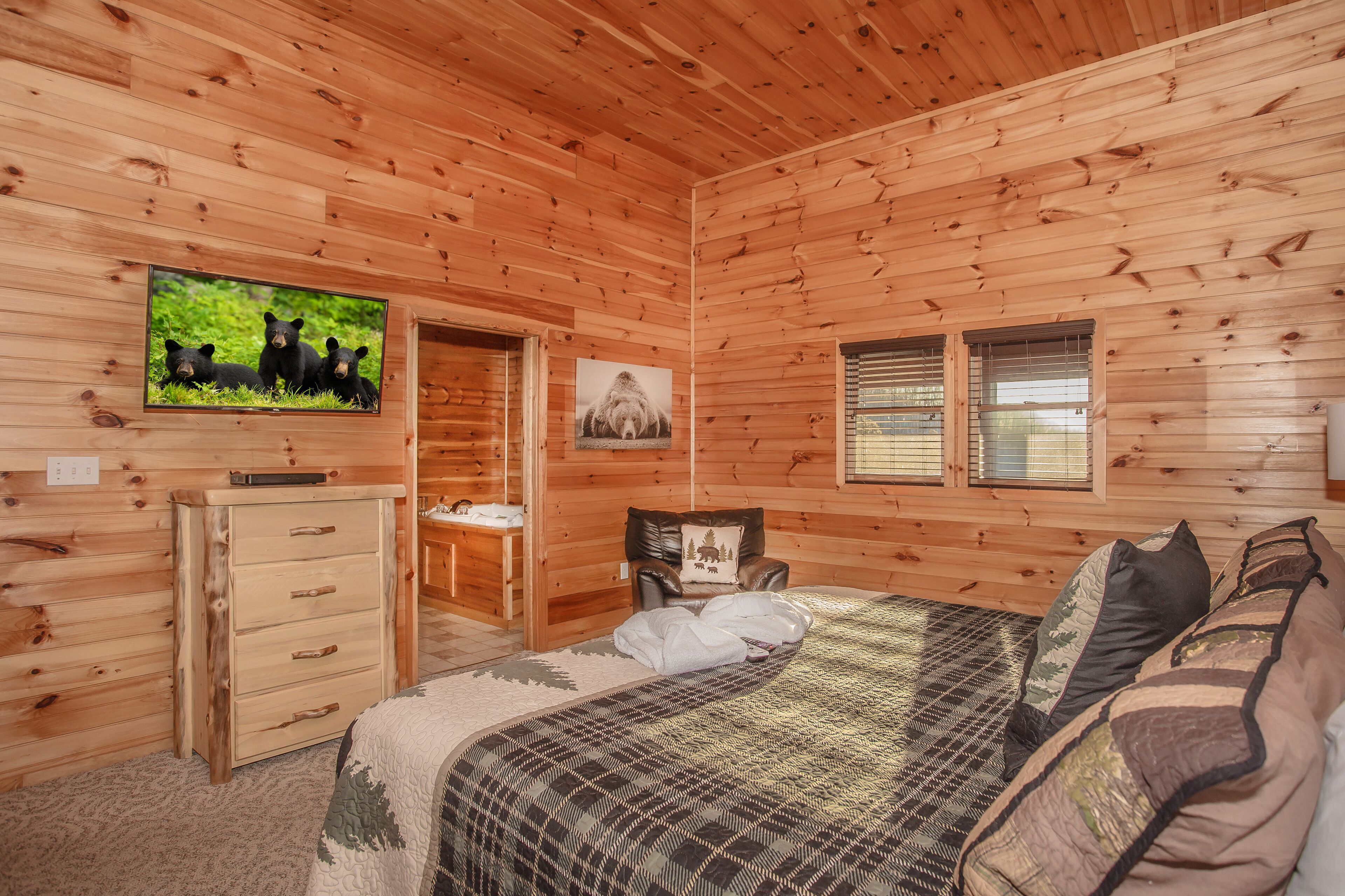 A wooden cabin bedroom with a bed, a dresser, a television, and a window.