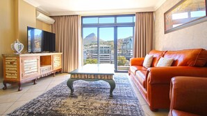 A living room with a television, a wooden cabinet, a coffee table, an orange sofa, and a large window with a city view. - Pristine Apartment in the Heart of Cape Town (Cape Town)