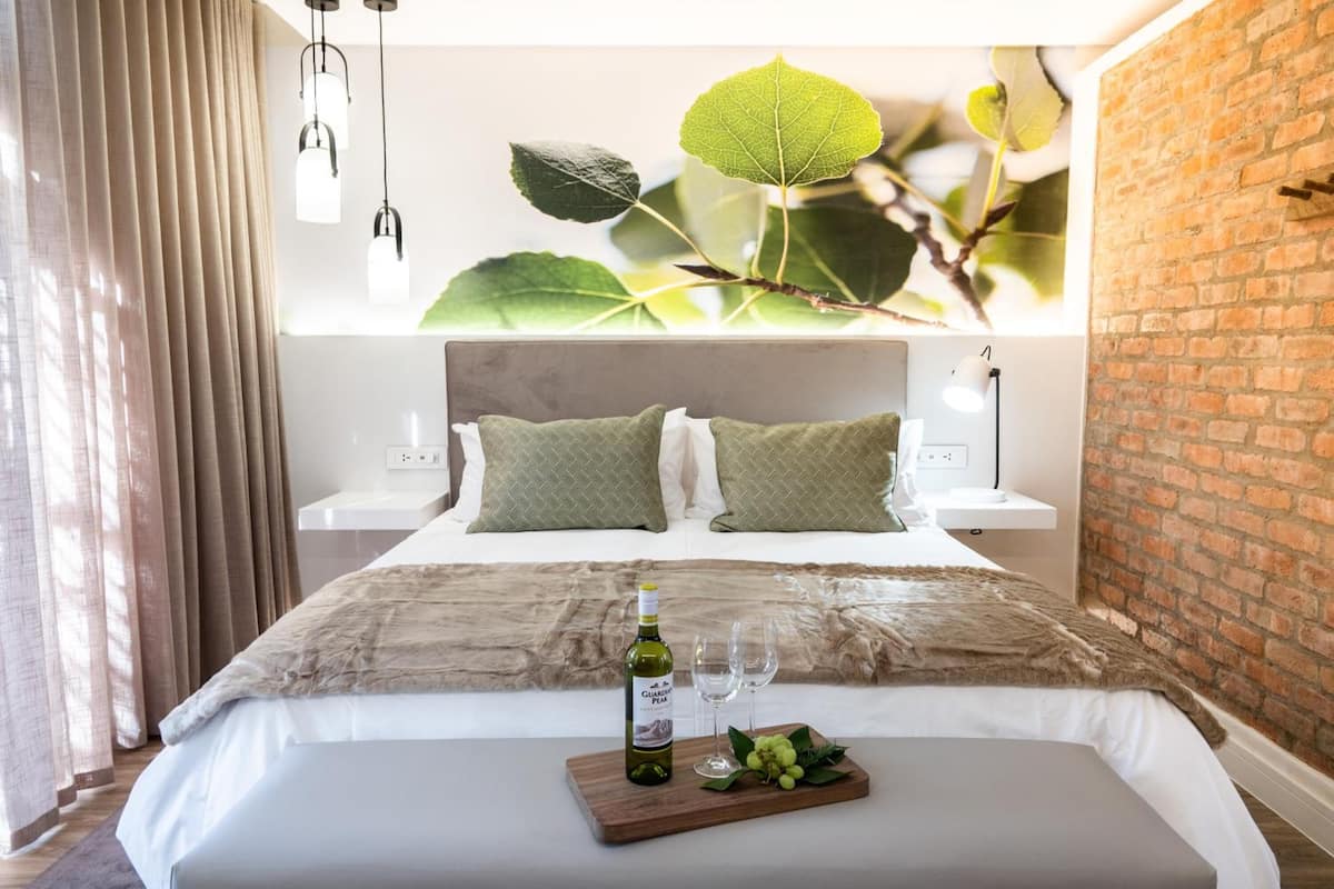 Ein modernes Schlafzimmer mit Backsteinwand, großem Fenster mit Blick auf grüne Blätter, einem Bett mit weißem Kopfteil, zwei Nachttischen mit Lampen sowie einem Nachttisch mit einer Weinflasche und Gläsern.