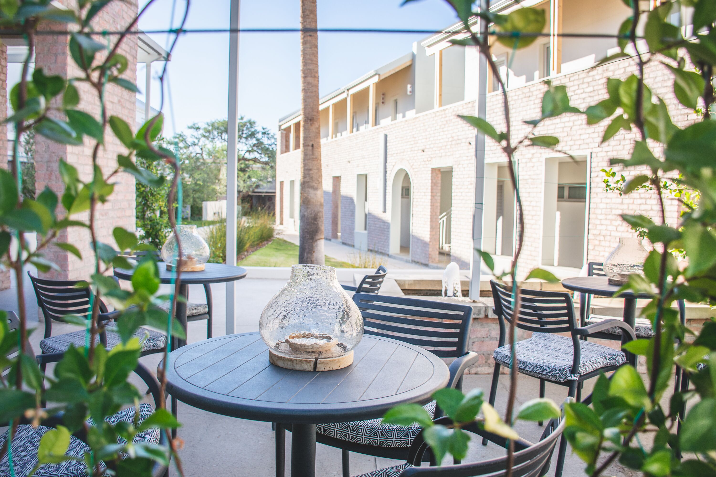 Un área de patio al aire libre con mesas y sillas redondas, un jarrón de cristal decorativo y un edificio de ladrillo al fondo.