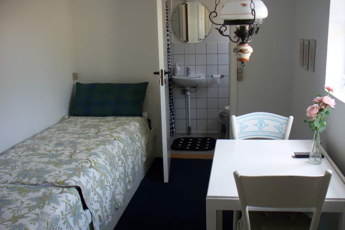 A bedroom with a bed, a bedside table, a chair, a small table, a lamp, a mirror, a sink, and a window.