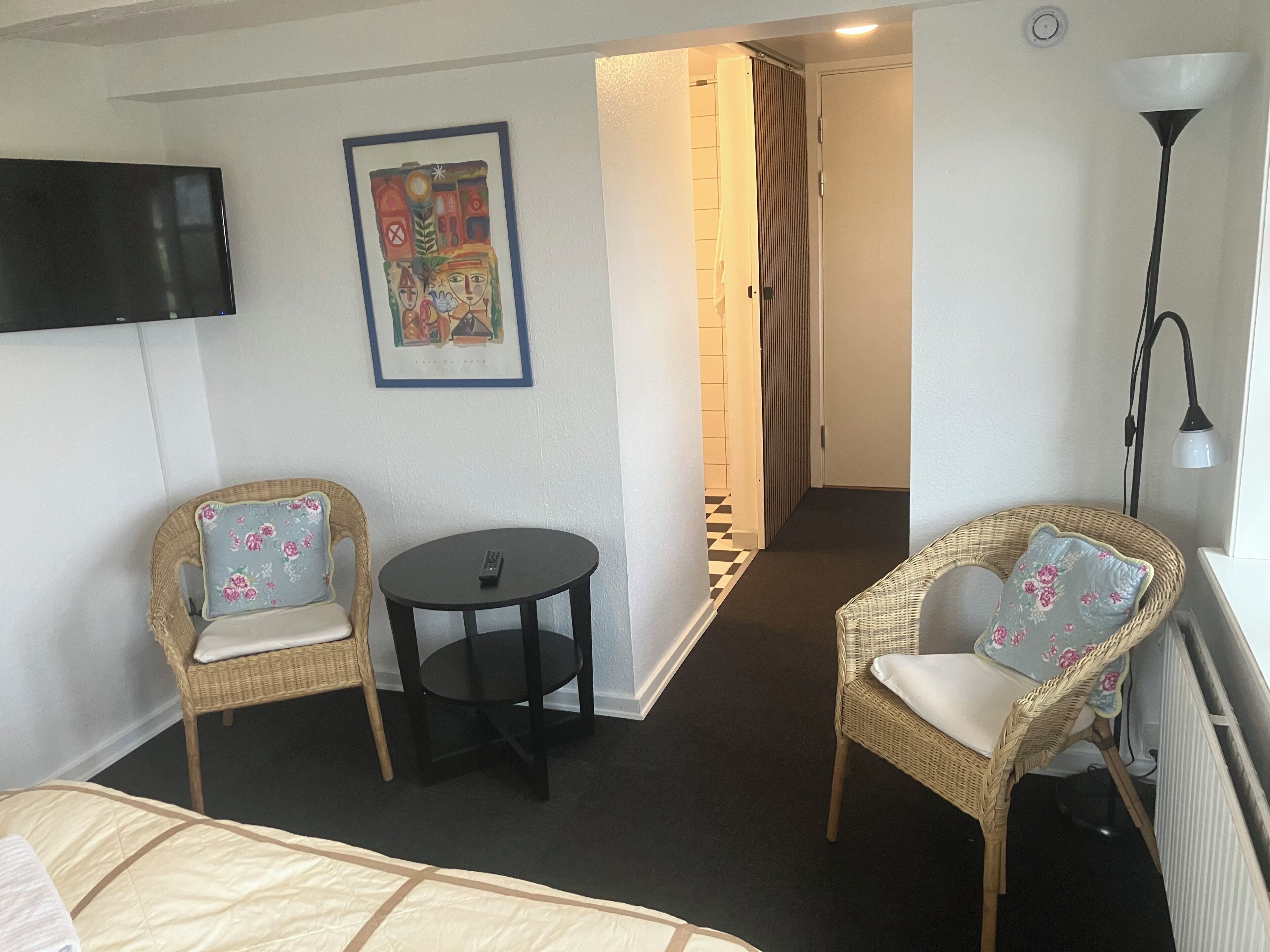 A hotel room with a bed, two wicker chairs with floral cushions, a small round table, a television mounted on the wall, and a framed artwork above the chairs.