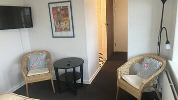 A hotel room with a bed, two wicker chairs with floral cushions, a small round table, a television mounted on the wall, and a framed artwork above the chairs.