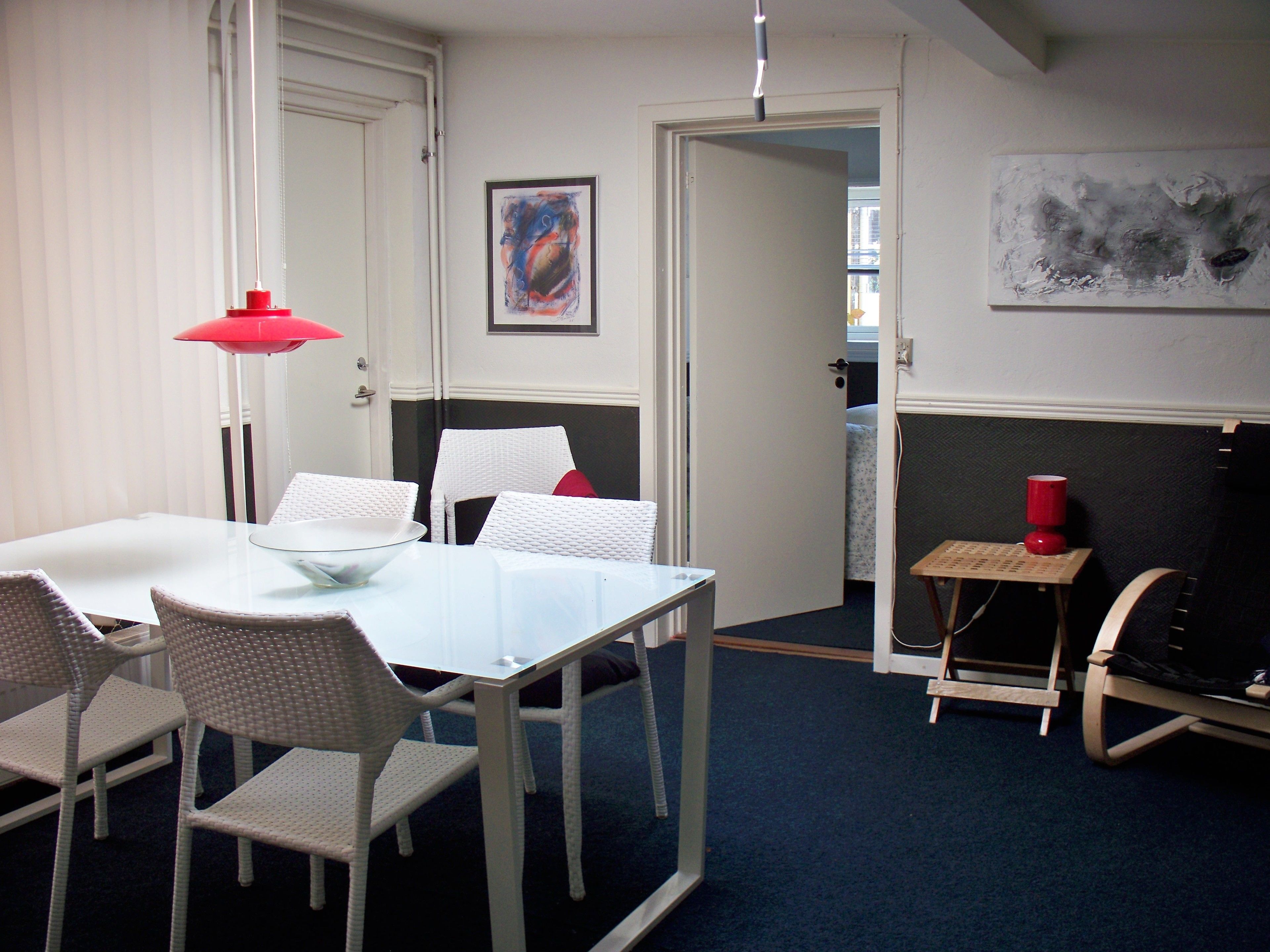 Una habitación con una mesa y sillas blancas, una lámpara roja, un taburete de madera y un cuadro en la pared.