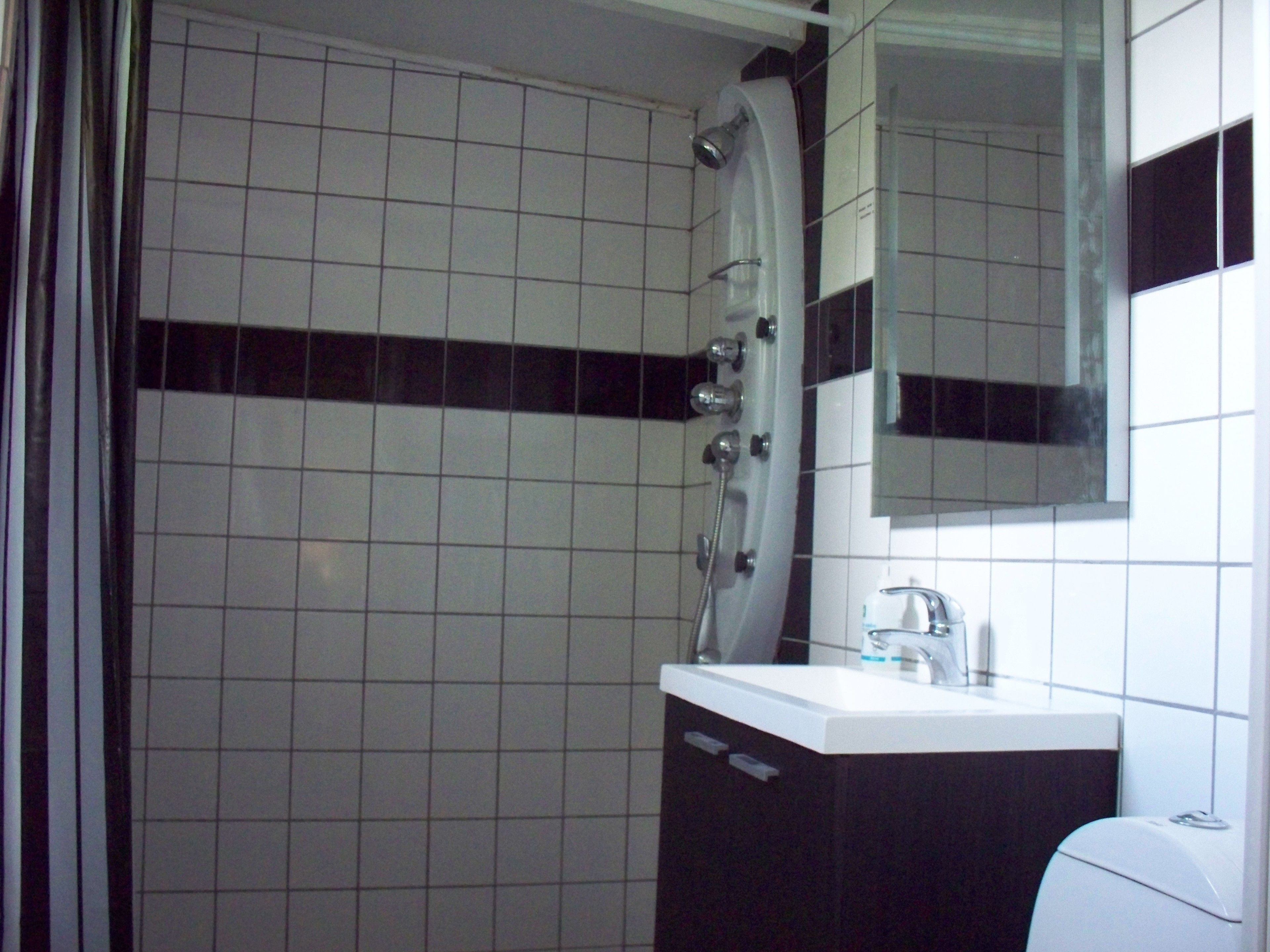 Une salle de bain avec des murs recouverts de carreaux blancs, une bande horizontale noire, un lavabo blanc avec un miroir au-dessus, et une douche au design incurvé.