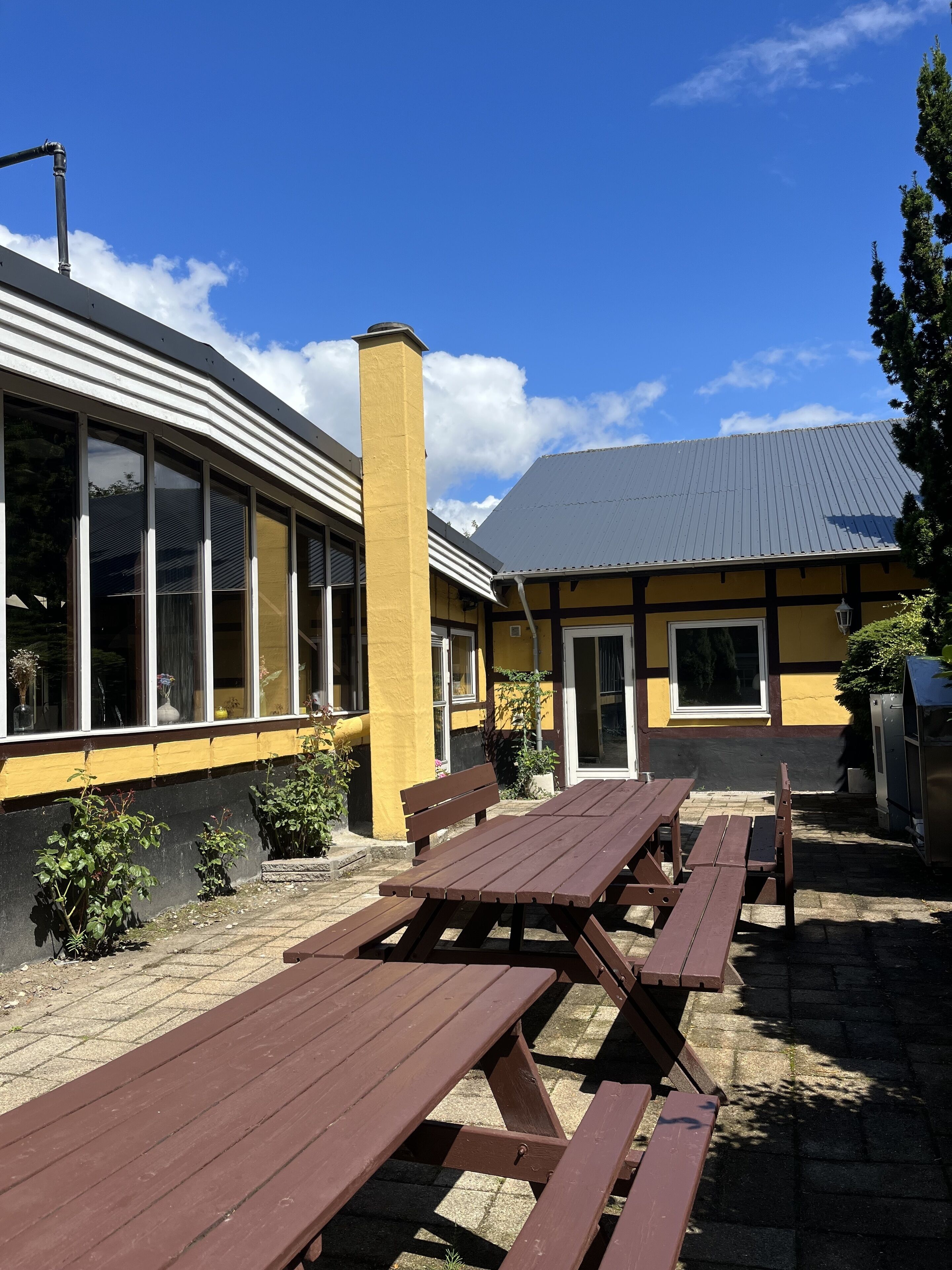 En utomhusplats med en uteplats, träbord och stolar för picknick, samt en gul byggnad med stora fönster.