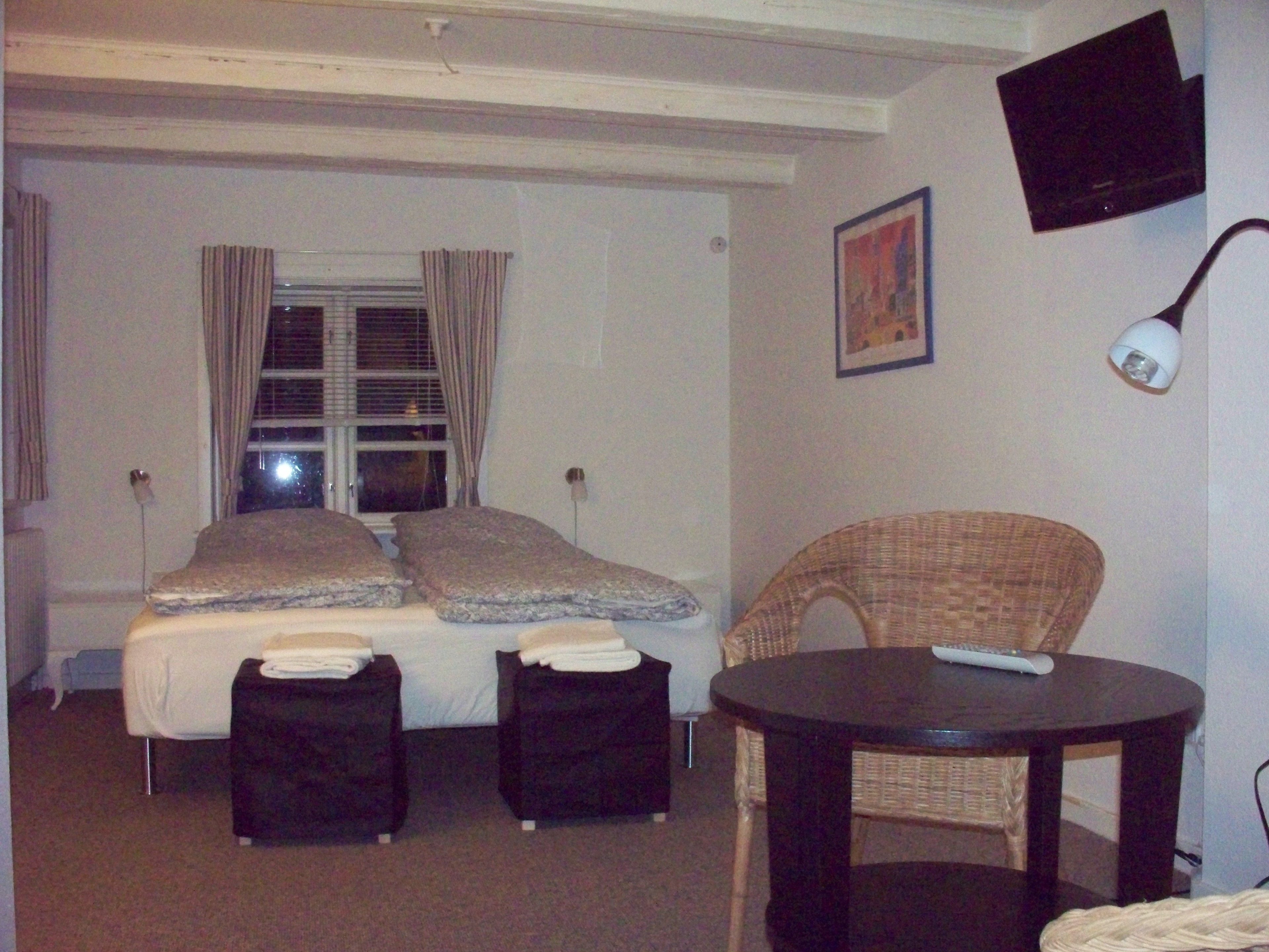 A hotel room with a bed, two bedside tables, a chair, a round table, a TV, and a window with curtains.