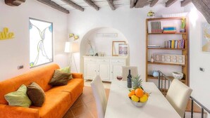 Una sala de estar acogedora con un sofá anaranjado brillante, una mesa de comedor blanca con fruta, y una estantería llena de libros y objetos decorativos.