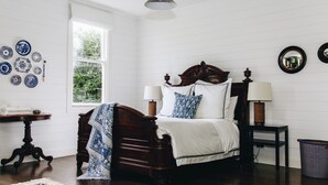 Une chambre avec un lit en bois, du linge de lit blanc, une table de chevet avec des lampes, une fenêtre avec des stores et un mur décoré d’assiettes.