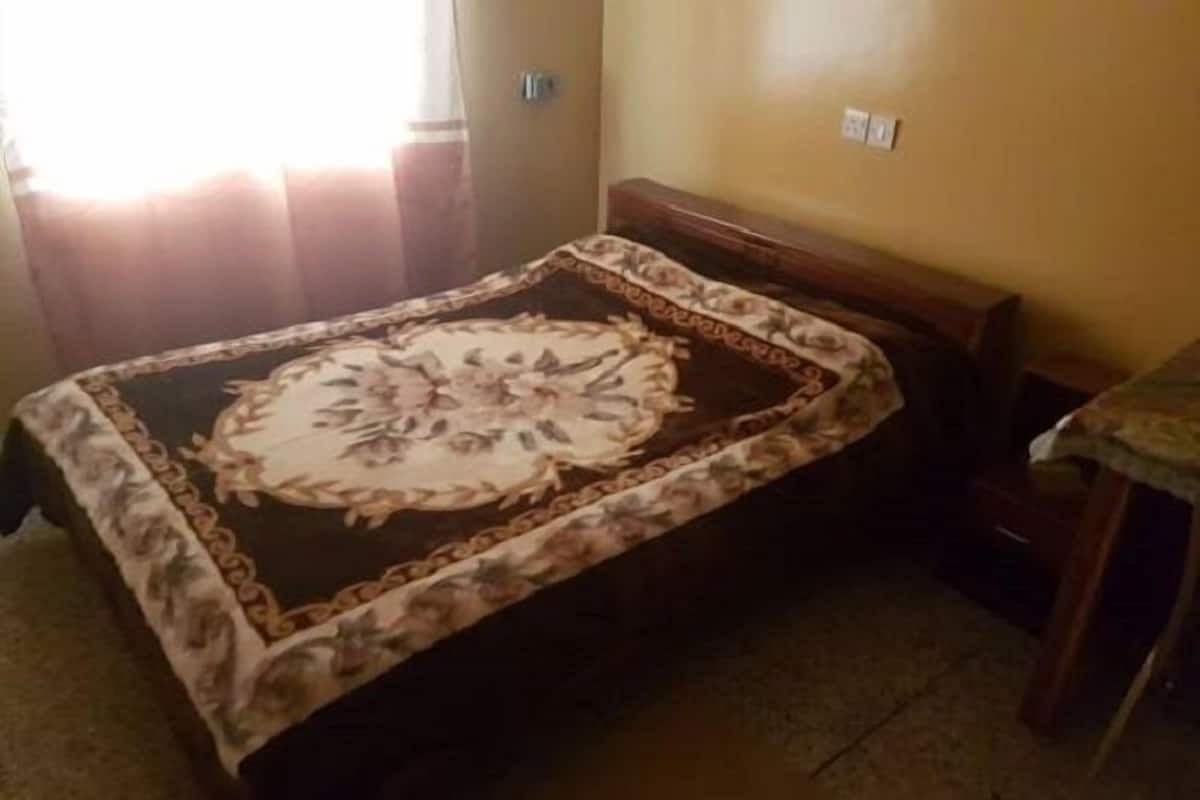 Une petite chambre avec un lit simple, une couvre-lit à motifs floraux, une tête de lit en bois et une petite table recouverte d’une nappe.