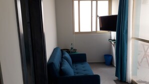 A small living area with a sofa, a television on a stand, a window with blinds, a water bottle, and a trash bin. - B&B Sereia Azul (Boa Vista)