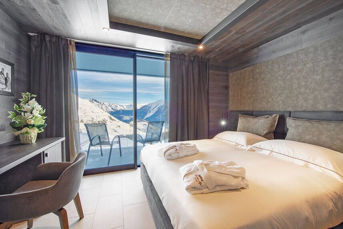 A modern hotel room with a large bed, a desk, and a view of mountains through a glass door.