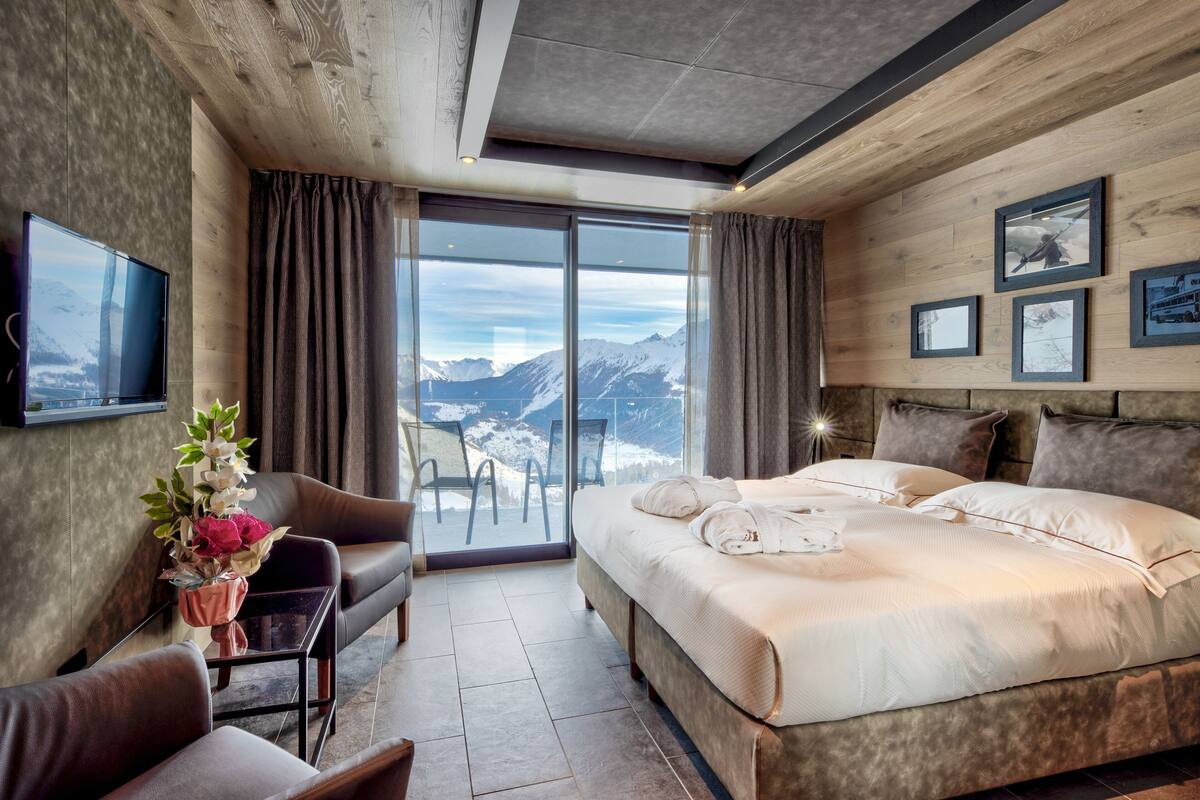 A hotel room with a large bed, a sofa, a small table with a vase of flowers, and a large window with a mountain view.
