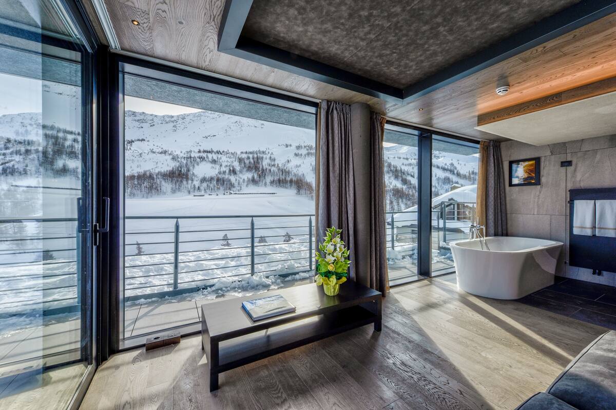 A modern bathroom with a freestanding bathtub, a large window with a snowy mountain view, and a wooden floor.