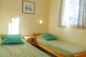 A room with two single beds, a small table, a wall-mounted lamp, and a window with curtains.