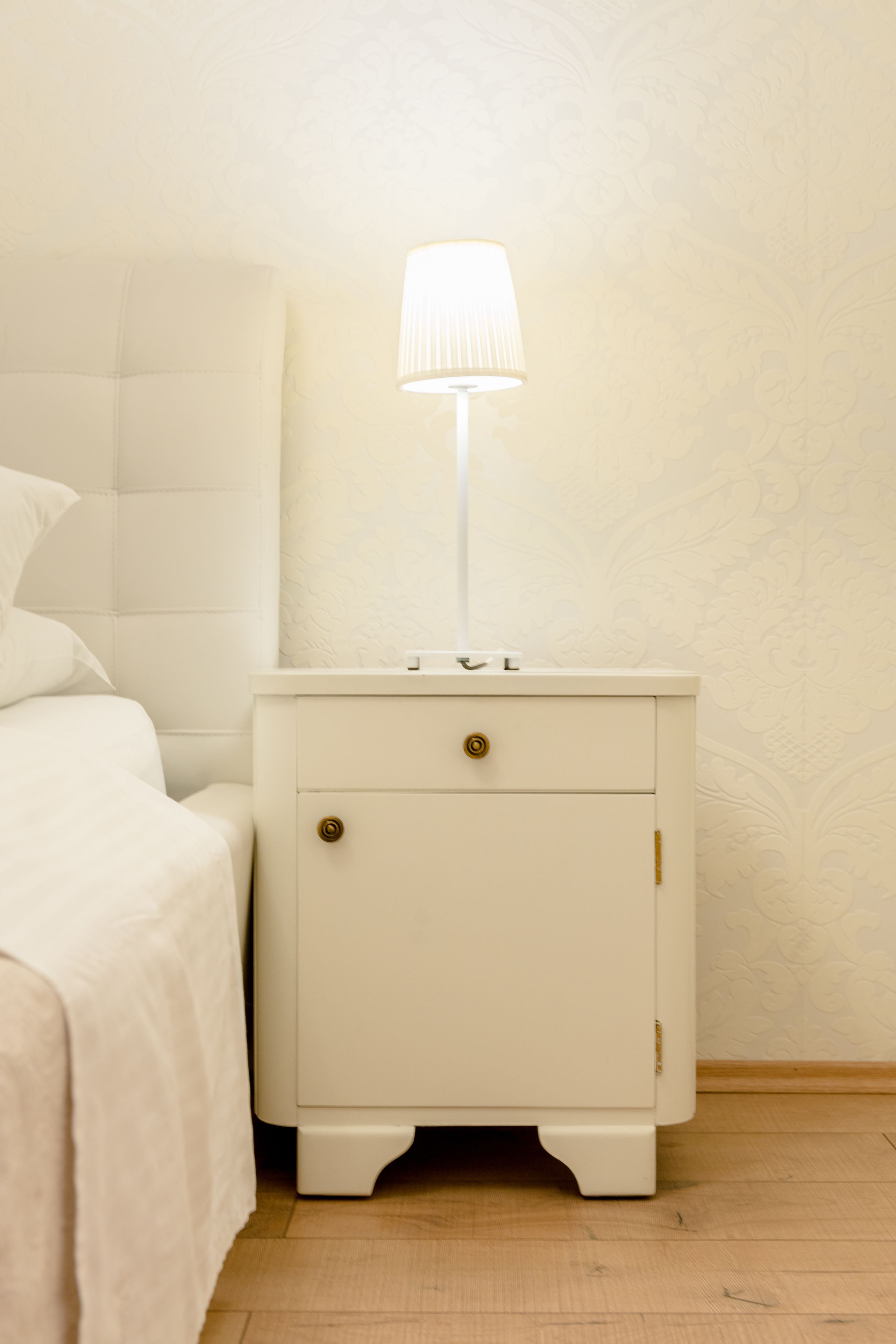 Una mesita de noche blanca con una lámpara, un cajón y una mesita auxiliar junto a una cama con cabecera.