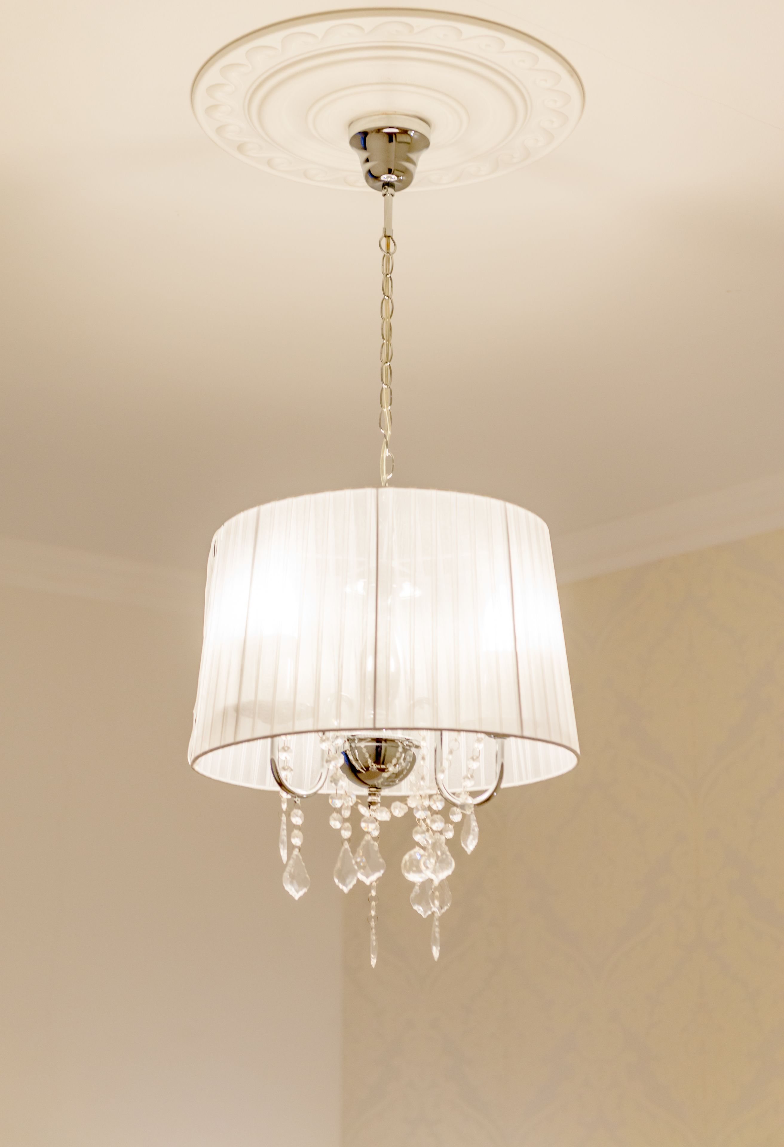 Un lampadari amb pantalla cilíndrica i detalls de cristall penjant d'un sostre amb una medalla decorativa.
