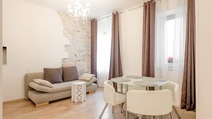 A modern living room with a stone wall, a glass dining table, white chairs, a sofa with cushions, and a small white side table. - Aurum Templi Suites (Split)
