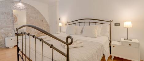 Quarto bem arrumado com estrutura de cama de metal, roupa de cama branca, duas mesas de cabeceira com abajures e um arco de parede de pedra.