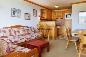 A cozy living room with a sofa and a small table, wooden kitchen cabinets, and a dining area with chairs.