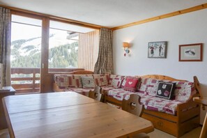 A cozy room with a wooden table, a sofa with patterned cushions, and a view of a mountainous landscape through a large window. - 10m from ski slopes, 3rd floor, balcony, 40m², Méribel-Mottaret (Méribel - Les Allues)