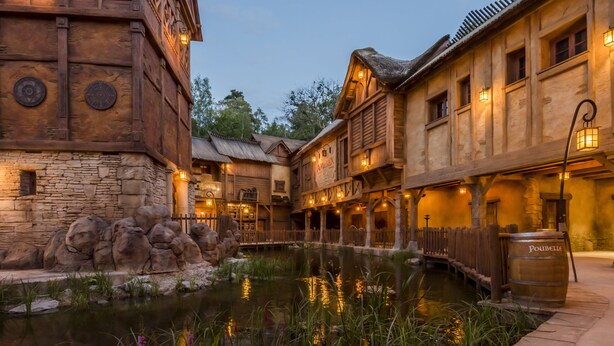 A rustic building with stone walls and wooden balconies, a small pond, and a wooden bridge. - Les Quais de Lutèce (Plailly)