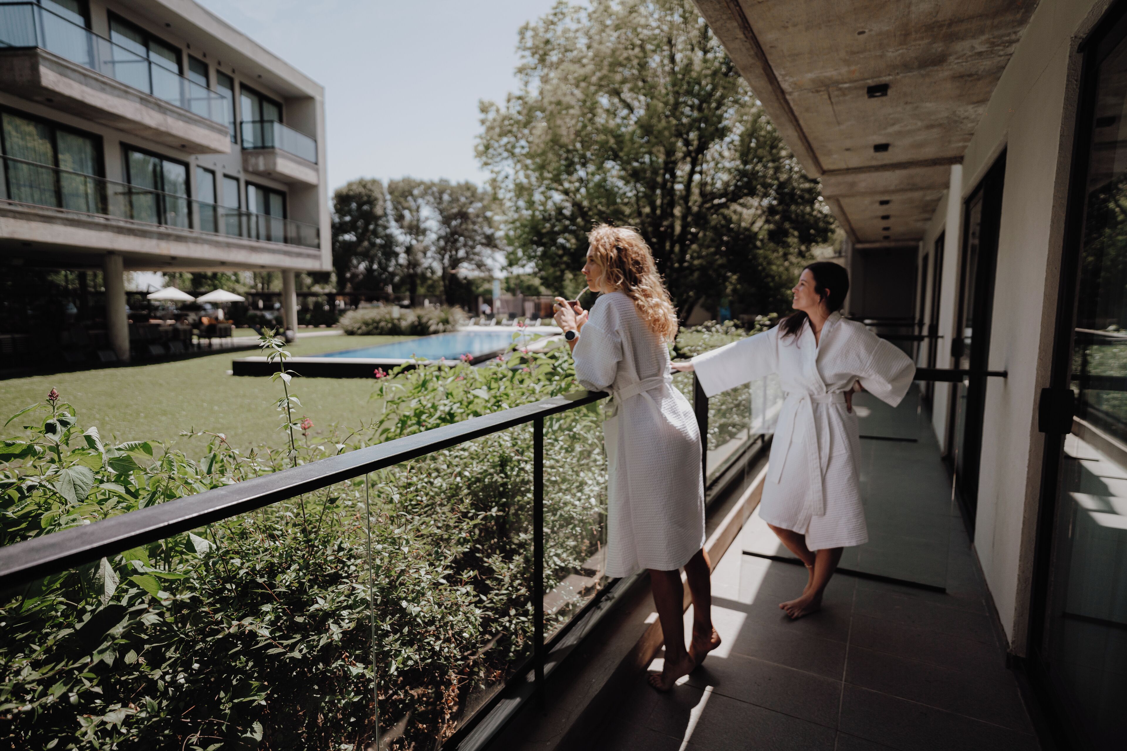 A balcony with a view of a swimming pool and greenery, a modern building in the background, and a person in a white robe.