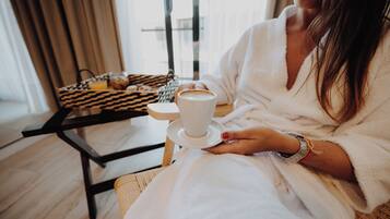 A person in a white bathrobe holding a cup of coffee, seated at a table with a tray containing a muffin and a glass of orange juice.