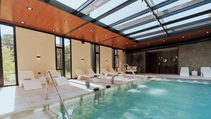 Una piscina interior moderna con techos de vidrio transparente, sillas de descanso y una pared de piedra.