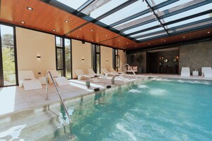 Una piscina interior moderna con techos de vidrio transparente, sillas de descanso y una pared de piedra.
