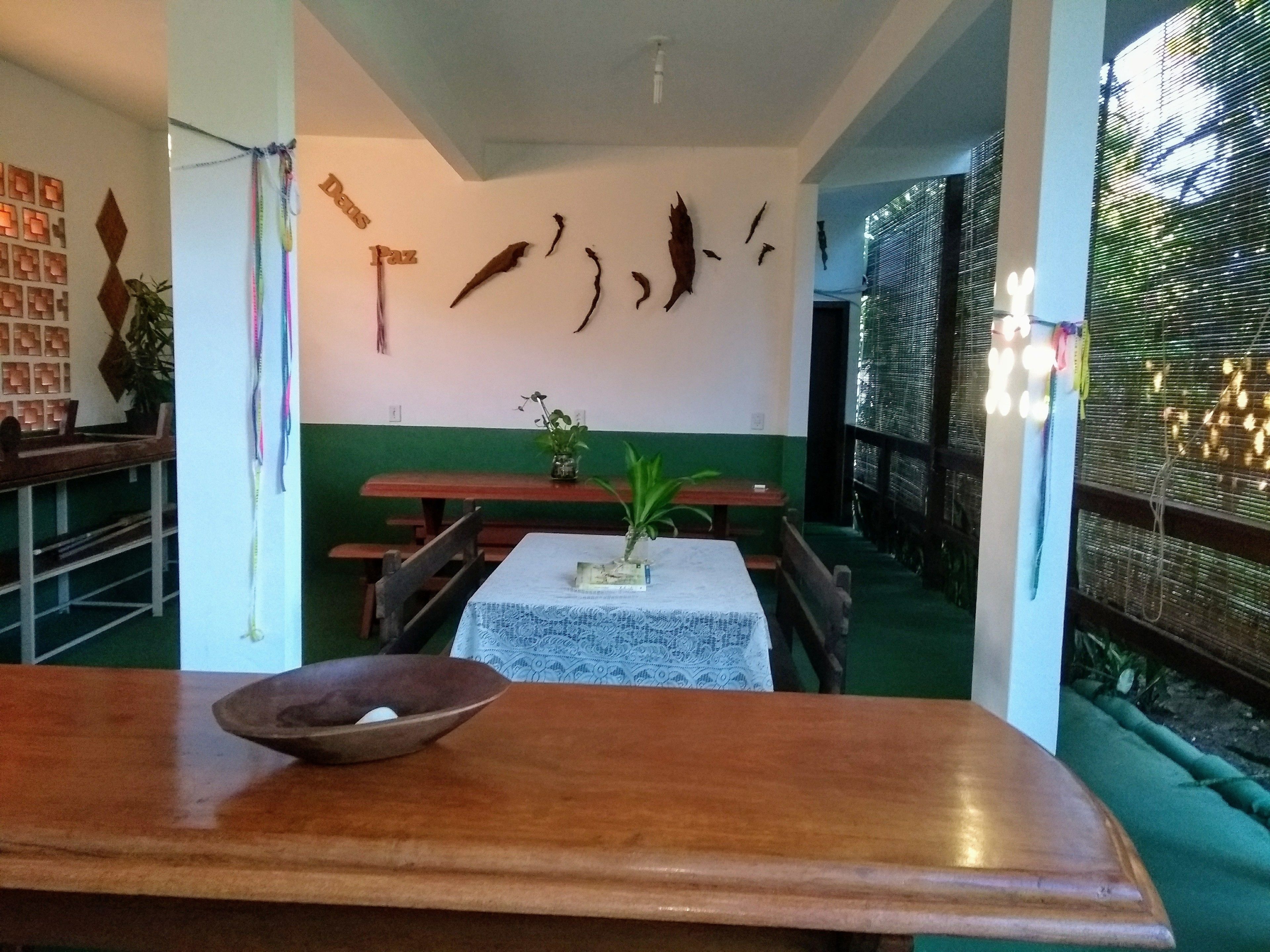 Uma área de refeições com mesas e bancos de madeira, uma parede decorativa com objetos pendurados e vista para o exterior através de uma janela com persianas.