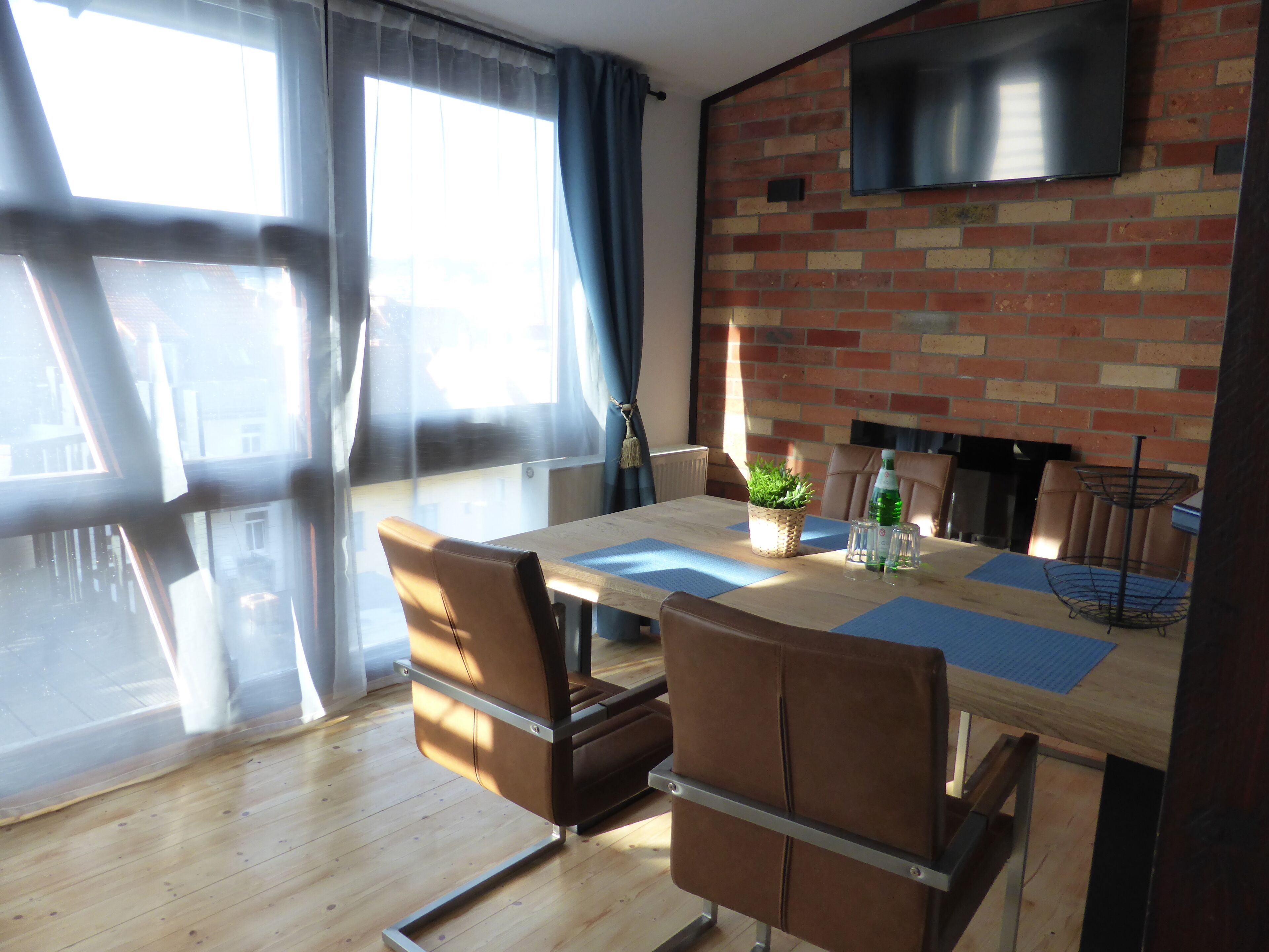 A modern dining area with a brick wall, a flat-screen TV, and a large window with curtains.