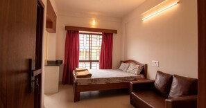 A bedroom with a bed, a wooden bench, a window with red curtains, and a wall-mounted light.